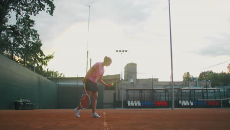 La-Chica-En-La-Línea-De-Servicio-Central-Golpea-Repetidamente-La-Pelota-En-La-Cancha,-Atrapa-La-Pelota-Con-La-Mano-Y-Sirve.-El-Plano-General.